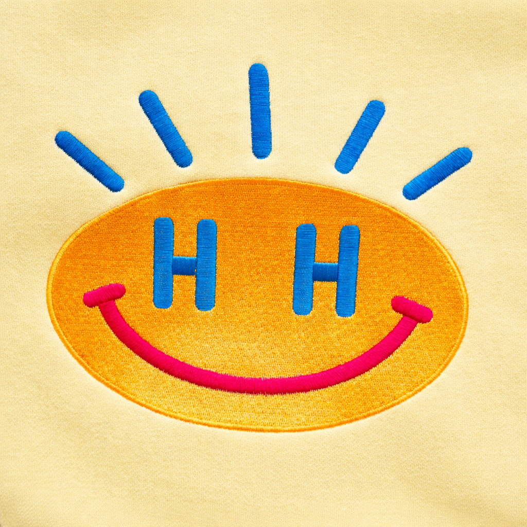 Close-up of Tatami embroidery SMILEY face on cool lemon yellow sweatshirt, showing flat woven stitching with subtle sheen.